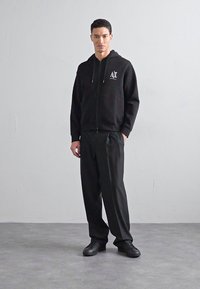 Juodas zip-up džemperis su logotipu, raukšlėti rankogaliai ir priekiniai kišeniai, derinamas su laisvomis juodomis kelnėmis ir juodais kedais ant pilko grindų.
