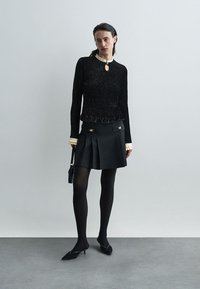 Pull noir texturé avec ourlet froncé, décolleté en forme de trou de serrure, associé à une jupe plissée noire avec des accents dorés. Collants brillants et chaussures à talons.