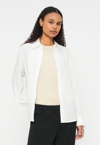 Bílá košile na knoflíky s límcem a dlouhými rukávy, nošená přes béžový top. Košile má volný střih a hladký povrch.