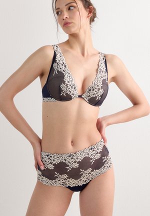 Woman wearing navy blue lingerie with white floral lace detailing, posing with one hand on hip and looking to the side against a plain background.