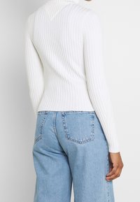 Pull à col roulé blanc côtelé avec manches longues, coupe ajustée, et détail en forme de V dans le dos, associé à un jean bleu clair.