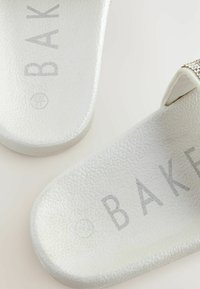 Baltas čības ar teksturētu zoli, ar pelēkiem uzdrukātiem burtiem "BAK" uz ieliktnēm. Gludas siksnas ar nelielu līkumu.