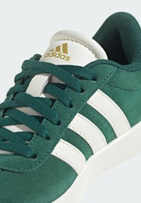 Zapatillas de ante verde con rayas de cuero blanco y cordones a juego. Logo dorado en la lengüeta. Tejido texturizado en el collar.