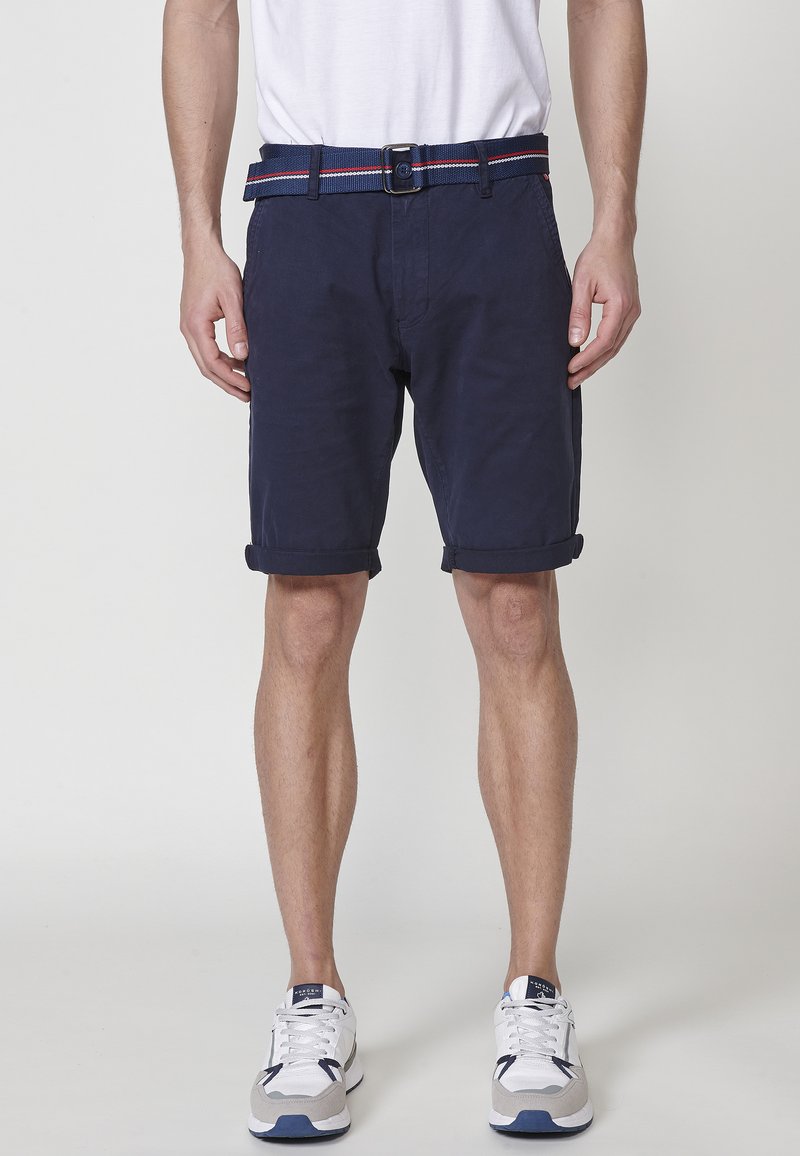 Man in marineblauwe opgerolde shorts, witte sneakers en een blauwe stoffen riem met rode en witte strepen, staande tegen een witte achtergrond.