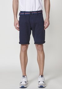 Homme portant un short bleu marine remonté, des baskets blanches et une ceinture en tissu bleue avec des rayures rouges et blanches, se tenant sur un fond blanc.