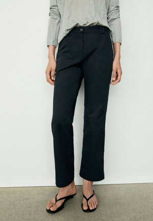 Personne portant un pantalon noir droit, une chemise grise à manches longues et des sandales noires, debout devant un fond blanc.