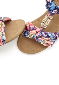 Hnedé ploché sandále majú opletený, kvetinový popruh z látky v ružovej a modrej farbe, doplnený o kovové detaily pre väčšiu eleganciu.