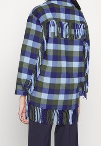 Ternet flanneljakke med blå, grønne og marineblå firkanter, frynser langs kanten og ryggen samt ærmer med knapper.