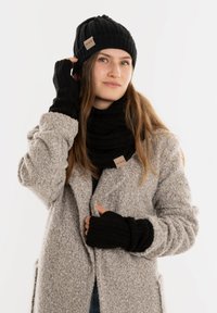 Zwarte gebreide beanie, sjaal en vingerloze handschoenen gedragen met een grijze gestructureerde jas. Kenmerken zijn ribpatronen en een klein label op de sjaal.