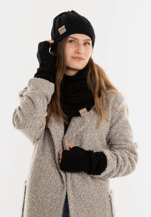 Zwarte gebreide beanie, sjaal en vingerloze handschoenen gedragen met een grijze gestructureerde jas. Kenmerken zijn ribpatronen en een klein label op de sjaal.