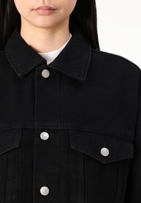 Schwarze Jeansjacke mit klassischem Design, silbernen Druckknöpfen und Brusttaschen mit spitzen Klappen. Weicher Stoff und robuste Verarbeitung.