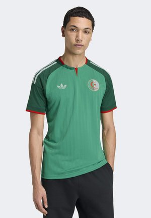 Joven con una camiseta de fútbol de Argelia verde con ribetes rojos, de pie con una mano en el bolsillo de sus pantalones negros, frente a un fondo claro.
