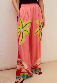 Un pantalon large rose présente des motifs de palmiers tropicaux en vert et en rose, avec des accents multicolores le long de l'ourlet, fabriqué dans un tissu doux.