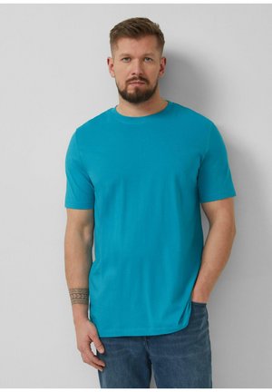 Man with beard and tattoo on left forearm wearing turquoise short-sleeve t-shirt and blue jeans, standing against a plain gray wall.