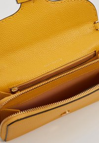 Cartera de cuero amarilla con un acabado texturizado, que cuenta con un compartimento con cremallera y un detalle de logo en el interior. Diseño minimalista.