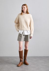 Beige, fluffy trui, gelaagd over een witte blouse, gecombineerd met grijze shorts en hoge bruine leren laarzen met ritsen en enkelriemen.