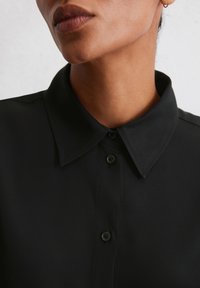 Schwarze Hemdbluse mit strukturiertem Kragen und dezenter Schimmerung. Verfügt über zwei schwarze Knöpfe an der Vorderseite und einen glatten Stofftextur.