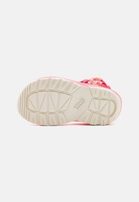 Una sandalia con suela texturizada beige y correa superior rosa, que presenta tejido trenzado y cordones elásticos ajustables, vista desde abajo.