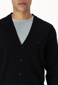Černý cardigan vyrobený z pleteného materiálu, s výstřihem do V, obsahující pět černých knoflíků a decentní černé logo Lacoste na levé straně.