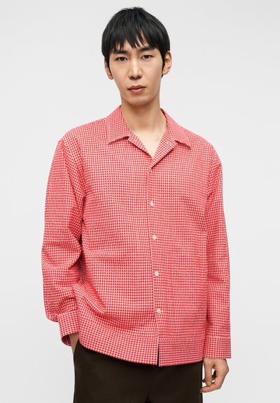 Νέος με κοντά μαύρα μαλλιά που φοράει κόκκινο και άσπρο πουκάμισο με κουμπιά με καρό μοτίβο και σκουρόχρωμο παντελόνι, μπροστά από μια απλή φόντο.