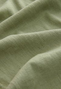 Tessuto di un verde chiaro, presentato in pieghe, caratterizzato da una trama morbida e naturale con fibre visibili e senza motivi o accenti distinti.
