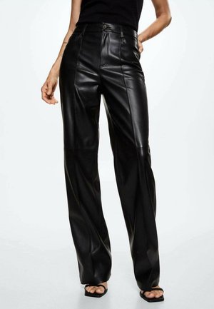 Vrouw draagt zwarte high-waisted leren broek met rechte pijpen, gecombineerd met zwarte open-toe sandalen met hak, staand tegen een effen achtergrond.