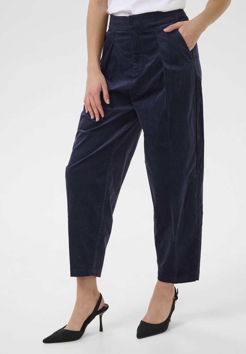 Pantaloni blu navy a vita alta con pieghe frontali e tasche laterali. Indossati con décolleté neri con tacco a scomparsa e punta a punta.