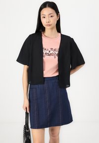 Svart öppen kortärmad cardigan över en rosa t-shirt med blommönster, kombinerad med en jeanstroppkjol med vertikala ränder.