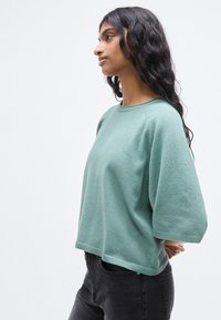 Sweatshirt oversize vert clair avec un col rond, des manches trois-quarts et un ourlet côtelé, associé à un jean foncé. Texture douce.