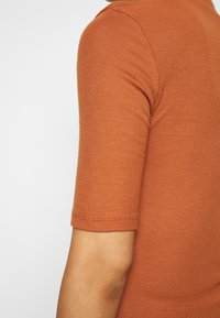 Primo piano del braccio superiore di una persona che indossa una maglietta a maniche corte, a coste, di colore arancio bruciato su uno sfondo bianco semplice.