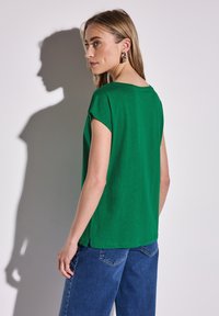 Vihreä lyhythihainen paita, jossa on pyöreä kaula-aukko ja sivuaukot, paritettuna sinisten farkkujen kanssa. Sileä rakenne, rento istuvuus.