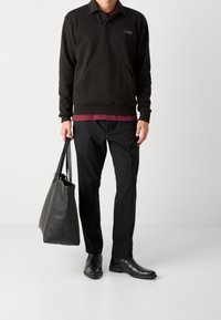 Černá mikina s kapsou, červená košile pod ní, černé kalhoty a černé kožené boty. Drží černou koženou tašku na rameno.