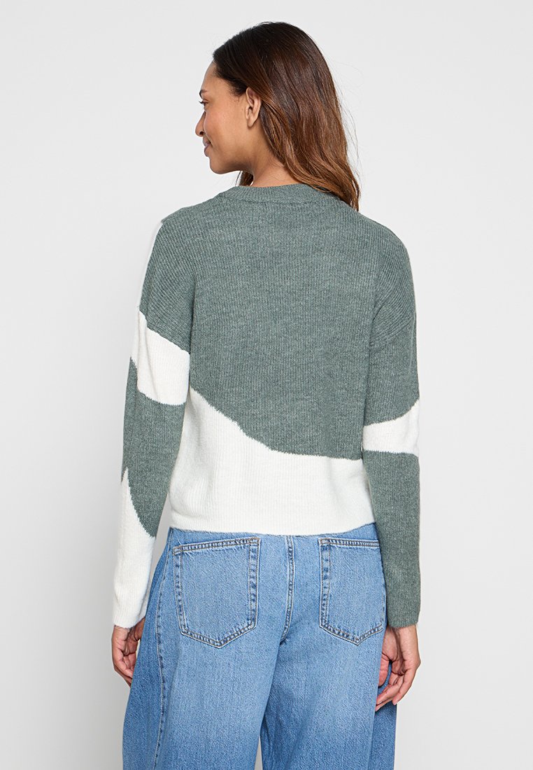 Grüner und weißer gestrickter Pullover mit Rundhalsausschnitt und abstraktem Muster, mit gerippter Textur und lässiger Passform, kombiniert mit hellblauen Jeans.