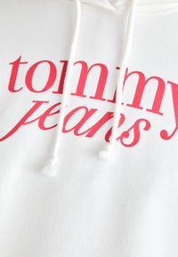 Vit hoodie med röd tryckt text "tommy jeans." Har en dragsko med vita tofsar och en jämn, mjuk textur.