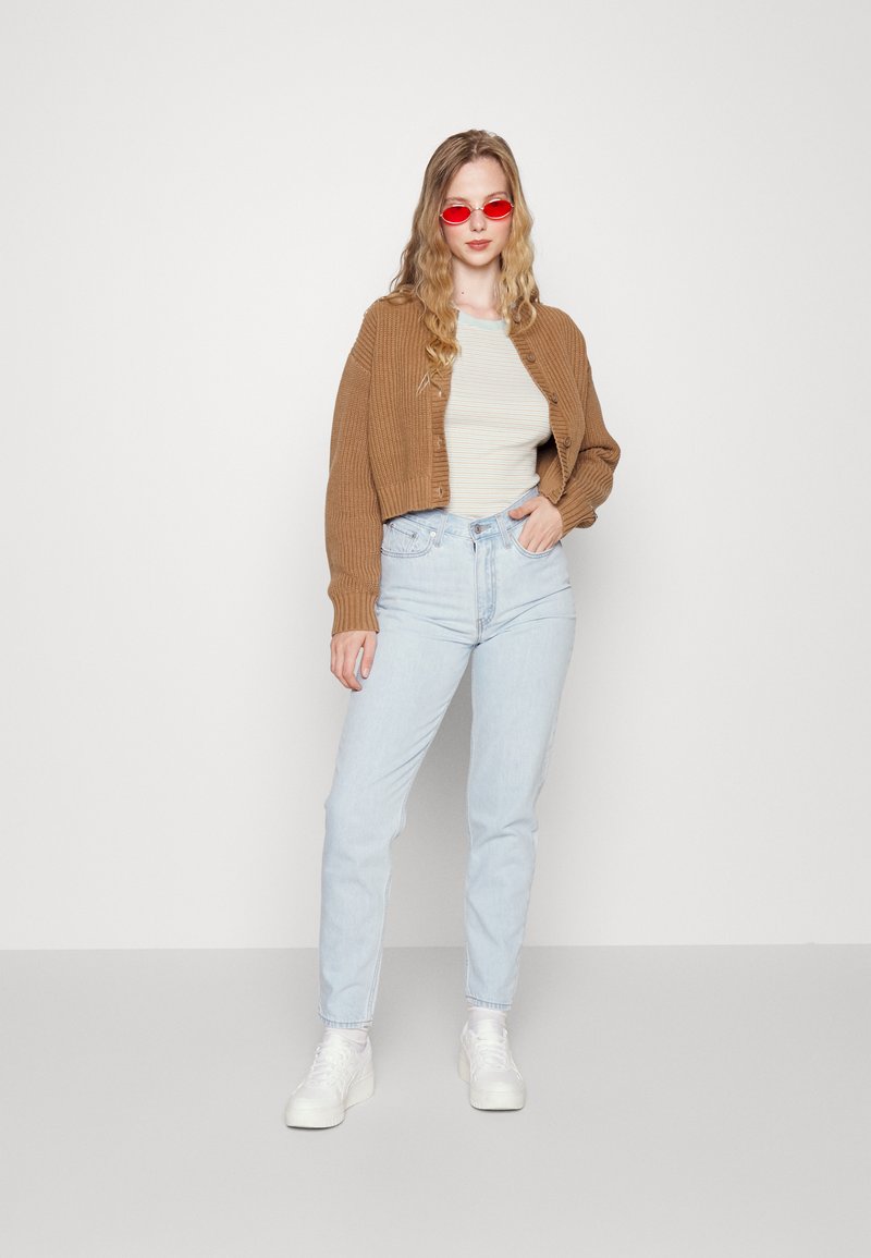 Brauner, verkürzter Cardigan, cremefarbenes, gestreiftes T-Shirt, hellblaue Jeans und weiße Sneakers. Das Model trägt rote Sonnenbrillen. Einfacher Hintergrund.