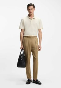 Beige Poloshirt, schmal zulaufende beige Hose, schwarze Lederhandtasche und schwarze Loafers. Einfaches Design mit dezenten Details und glatten Texturen.