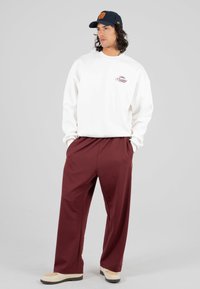 Uomo in piedi con le mani nelle tasche, indossa una felpa bianca, pantaloni bordeaux, sneakers crema e un cappellino blu navy con un logo arancione.