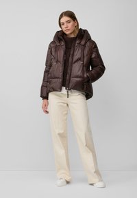 Braune gesteppte Jacke mit Glanz, Kapuze und Reißverschlüssen, über einem dunklen Strickpullover getragen. Kombiniert mit cremefarbenen Hosen mit weitem Bein und weißen Sneakers.