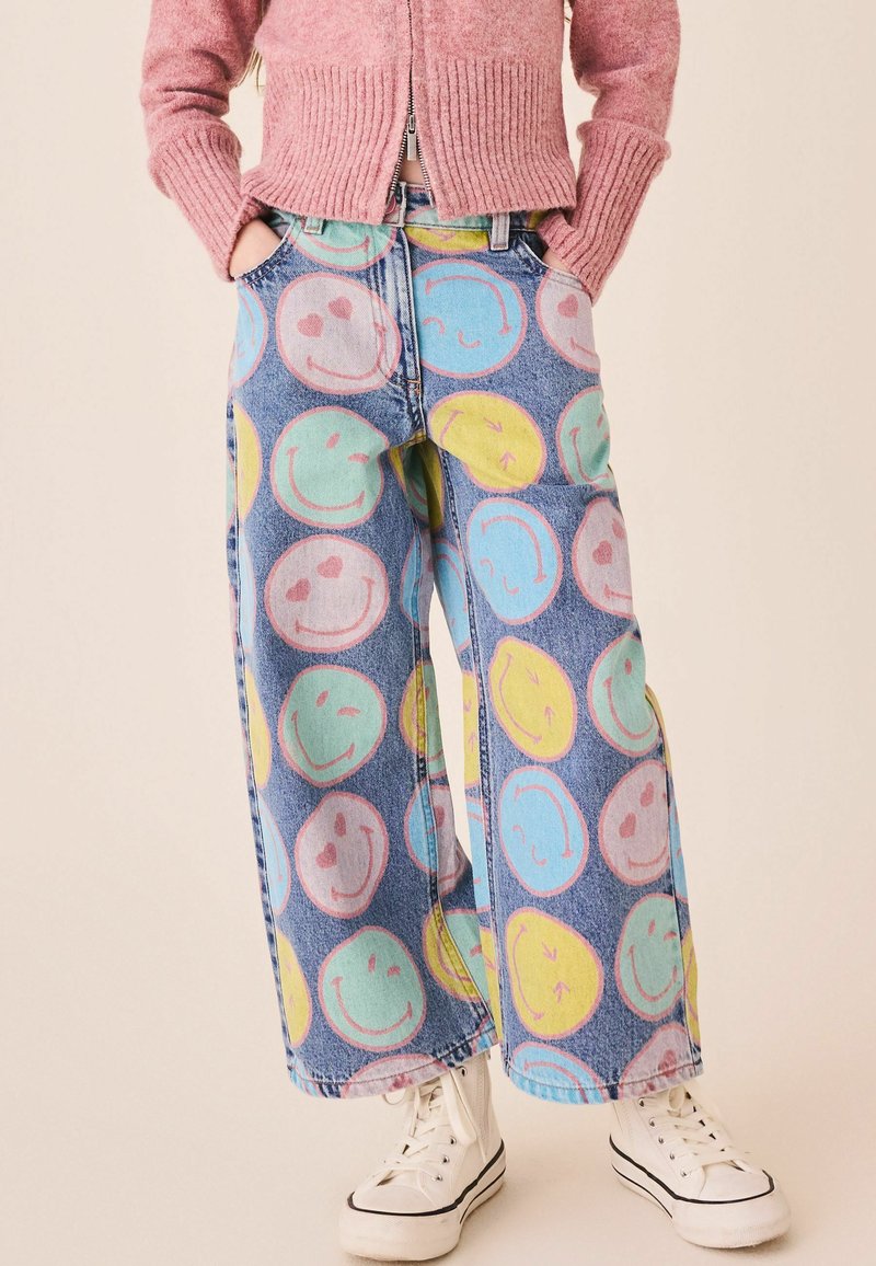 Pantalon en jean à jambes larges avec de grands imprimés colorés de visages souriants, porté avec un pull zippé rose et des baskets blanches montantes.