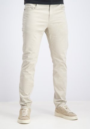 Man draagt strakke beige broek en lichtbeige suède sneakers, staand tegen een effen witte achtergrond.