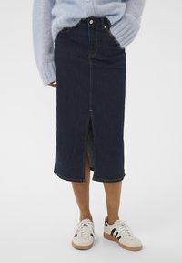 Dunkelblaue Denim-Midirock mit seitlichem Schlitz, heller kontrastierender Naht und tailliertem Bund. Kombiniert mit weißen Sneakers.