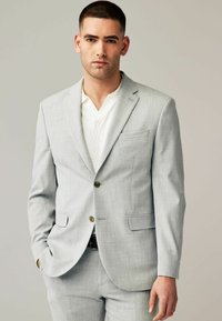 Traje gris claro con una chaqueta ajustada que presenta dos botones y bolsillos, combinada con una camisa blanca texturizada. Tela suave, diseño a medida.