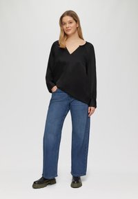 Zwarte zijden blouse met een v-hals en lange mouwen, gecombineerd met blauwe wijde jeans en zwarte chunky laarzen. Neutrale achtergrond.