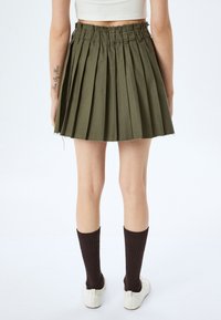 Olijfgroene geplooide rok met een rafelige zoom, voorzien van een gathered tailleband. Model draagt bruine sokken en witte schoenen, benen zichtbaar van achter.