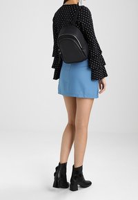 Svart faux läder ryggsäck med böjd design och dragkedja, bärs över en långärmad svart blus med vita prickar och en blå kjol.