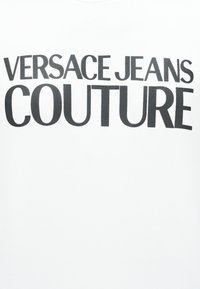 Μαύρο κείμενο «VERSACE JEANS COUTURE» με κεφαλαία γράμματα σε λευκό φόντο, με τη λέξη «COUTURE» σε μεγαλύτερη γραμματοσειρά κάτω από το όνομα του εμπορικού σήματος.