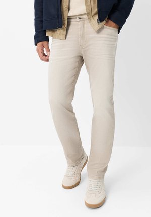 Mann trägt hellbeige Slim-Fit-Jeans, weiße Sneaker mit Gummisohlen und ein geschichtetes Outfit, bestehend aus einem beigen Hemd und einer dunklen Jacke.