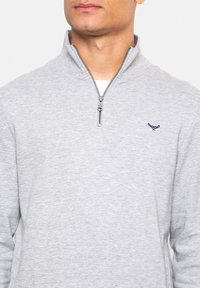 Pull léger gris clair avec fermeture éclair quart, fabriqué en tissu doux. Présente un logo bleu marine sur la poitrine et une finition texturée au niveau du col.