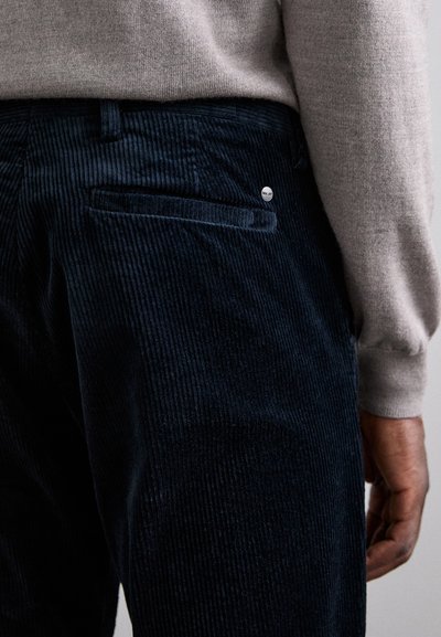 Navy corduroy pants with textured stripes, featuring a back pocket and a round metal logo accent. Styled with a light gray sweater.