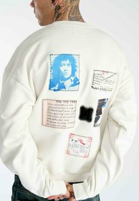 Weißer Hoodie mit Textil-Patches und grafischen Drucken, darunter ein blauer Porträt, Text und kontrastierende Texturen auf der Rückseite.